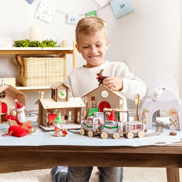 The BEST Advent Calendars for 2025 - 17 A young boy is playing with a wooden train set and miniature houses, imagining them as part of magical advent calendars, on a table in a brightly lit room.