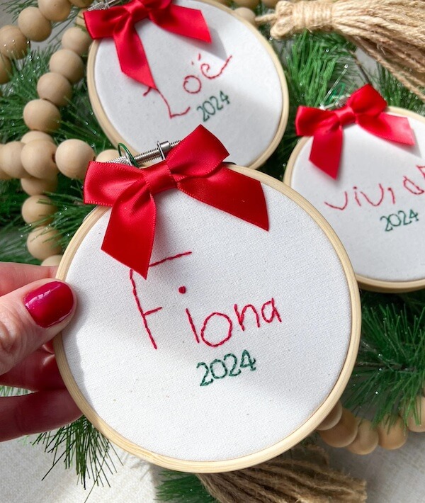 14 Southern Ornament & Stocking Shops We Love - 18 Three embroidery hoops with white fabric, each featuring a name and "2024" stitched in red and green thread, decorated with red bows. A charming example of Southern Christmas Decor, accented by greenery and beads in the background.