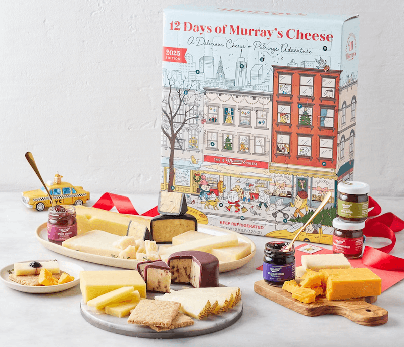 The BEST Advent Calendars for 2025 - 8 A 2025 cheese Advent Calendar box is surrounded by three plates of assorted cheeses, cheese knives, crackers, and jars of jam on a white surface.