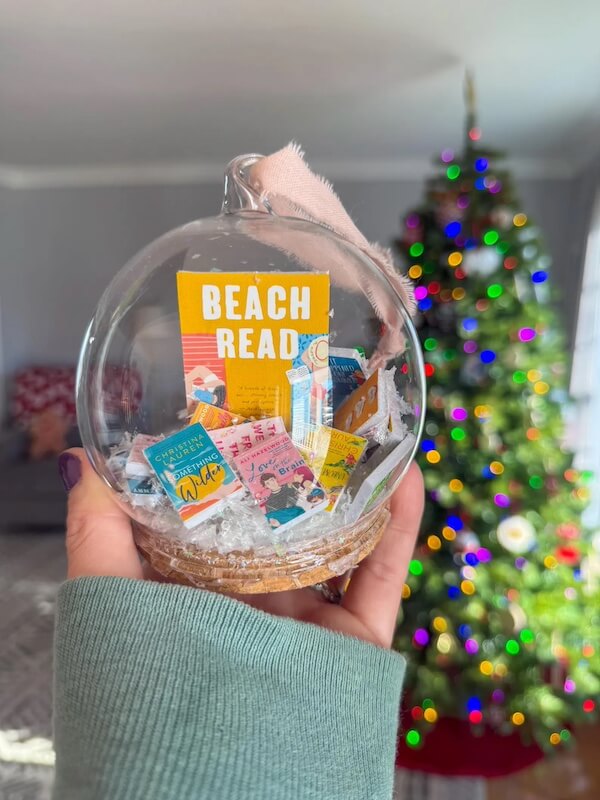 14 Southern Ornament & Stocking Shops We Love - 17 A hand holds a clear Southern ornament filled with mini book covers and artificial snow, with a decorated Christmas tree in the blurry background.