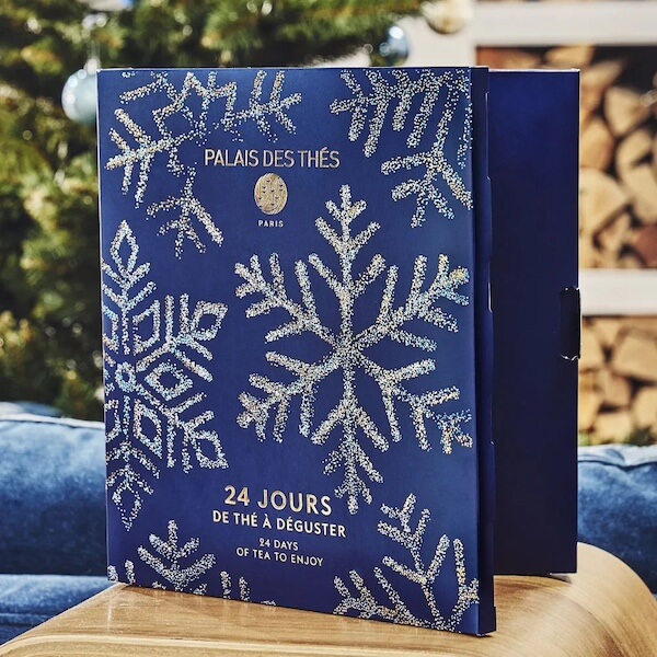 The BEST Advent Calendars for 2025 - 9 A blue Palais des Thés Advent Calendar with silver snowflake designs sits on a table, with a decorated Christmas tree and stacked firewood in the background, ready to bring festive cheer for 2025.