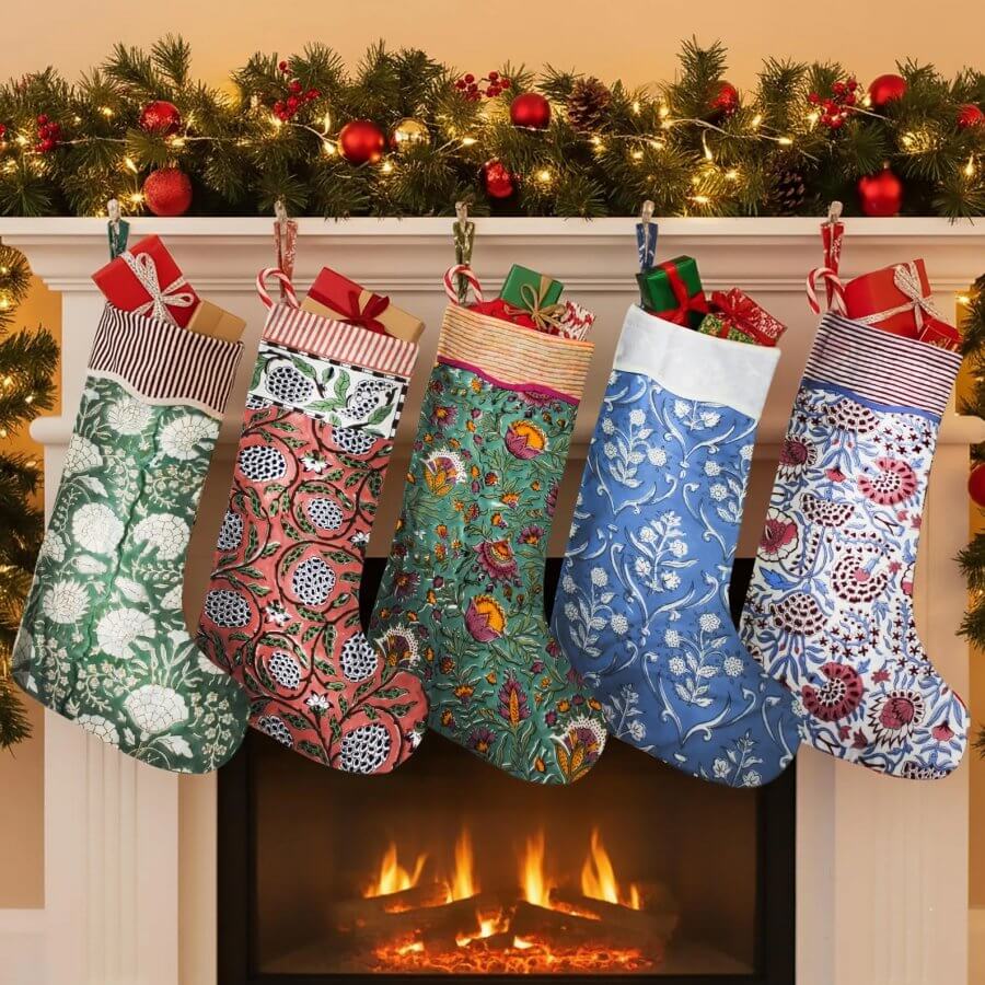 14 Southern Ornament & Stocking Shops We Love - 19 Five patterned Christmas stockings from local stocking purveyors hang from a decorated mantel above a lit fireplace, with garland and Southern Christmas decorations, including red ornaments, overhead.