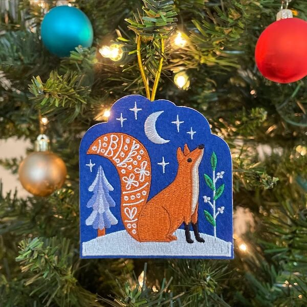 14 Southern Ornament & Stocking Shops We Love - 15 Embroidered ornament of a fox sitting under the moon, hanging on a Christmas tree with blue, gold, and red baublesβa charming touch of Southern holiday decor.