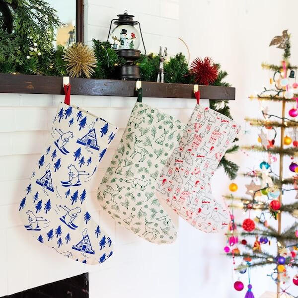 14 Southern Ornament & Stocking Shops We Love - 14 Three patterned Christmas stockings from local stocking purveyors hang from a decorated mantel above the fireplace, with a lit Christmas tree glowing on the rightβradiating classic charm inspired by Southern Christmas decorations.