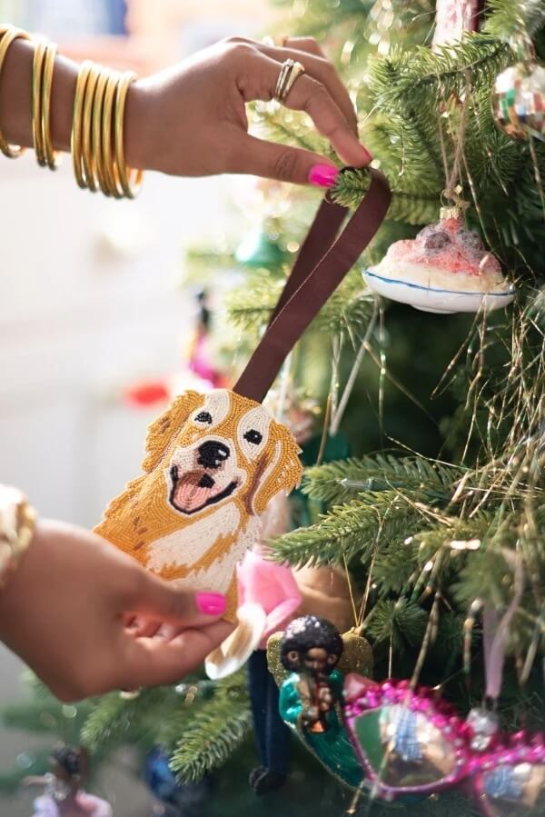 14 Southern Ornament & Stocking Shops We Love - 20 A person hangs a dog-shaped ornament with a ribbon on a Christmas tree decorated with colorful ornaments, adding a touch of charm inspired by Southern Ornament Shops.