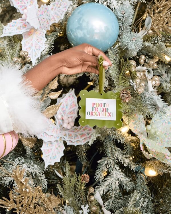14 Southern Ornament & Stocking Shops We Love - 16 A hand in a white fur-trimmed sleeve hangs a green "Photo Frame Ornament," a charming Southern ornament, on a decorated Christmas tree with ornaments and holly leaves.