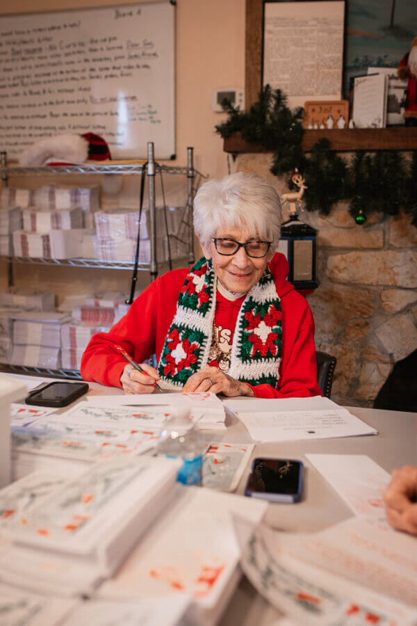 Meet the 94-Year-Old 'Chief Elf' Keeping the Magic of Christmas Alive - 2 An elderly woman in a festive scarf sits at a table, writing on papers. Stacks of mail and holiday decorations fill the background with seasonal cheer.