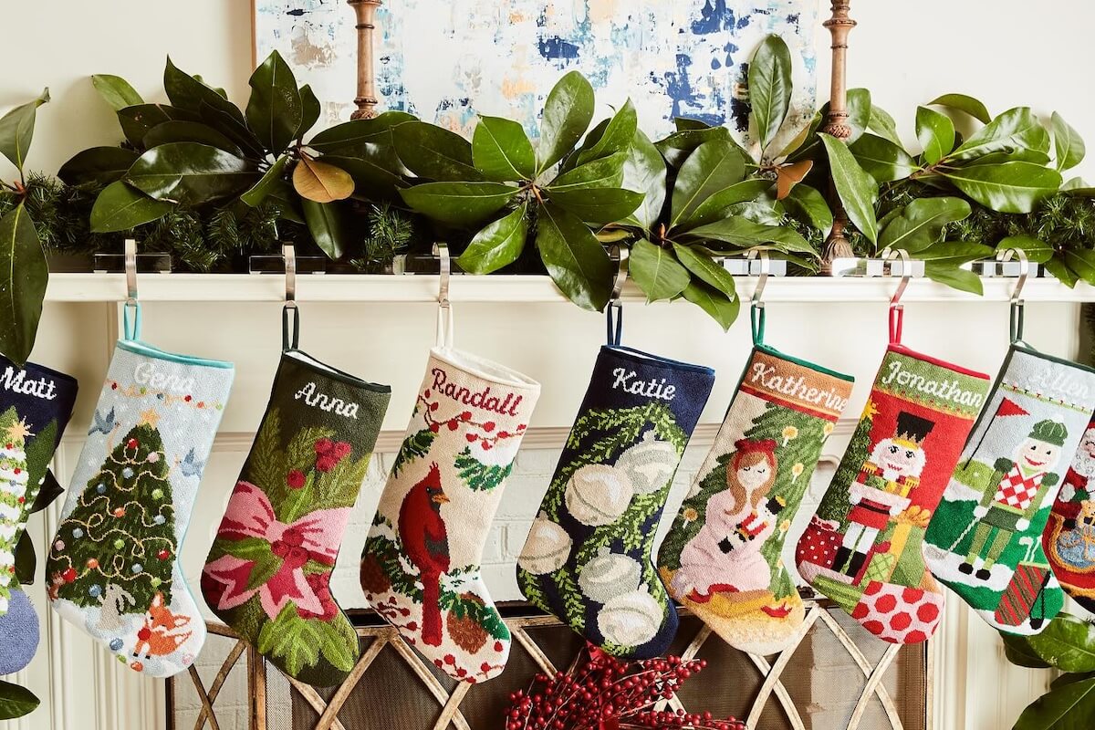 Seven colorful Southern Stockings with different designs and embroidered names hang from a decorated mantel with greenery, magnolia leaves, and a charming Southern ornament accent.