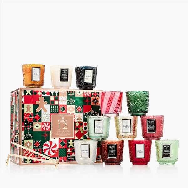 The BEST Advent Calendars for 2025 - 3 A set of twelve assorted decorative candles in colorful glass holders, arranged around a festive patterned box—perfect for gifting or pairing with advent calendars; a gold candle accessory is included.