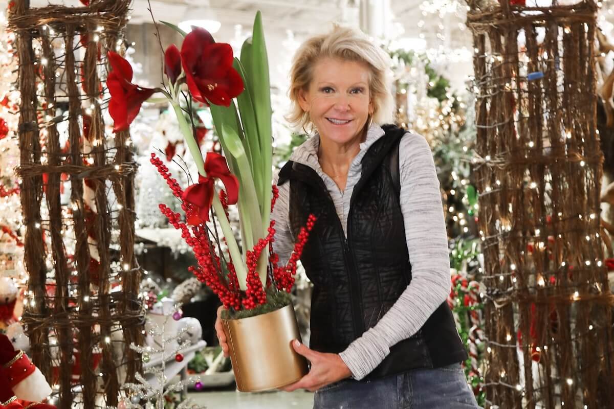 A woman stands indoors at Lucy's Market, holding a gold pot containing red flowers and berries, surrounded by festive holiday decorations and twinkling string lights.