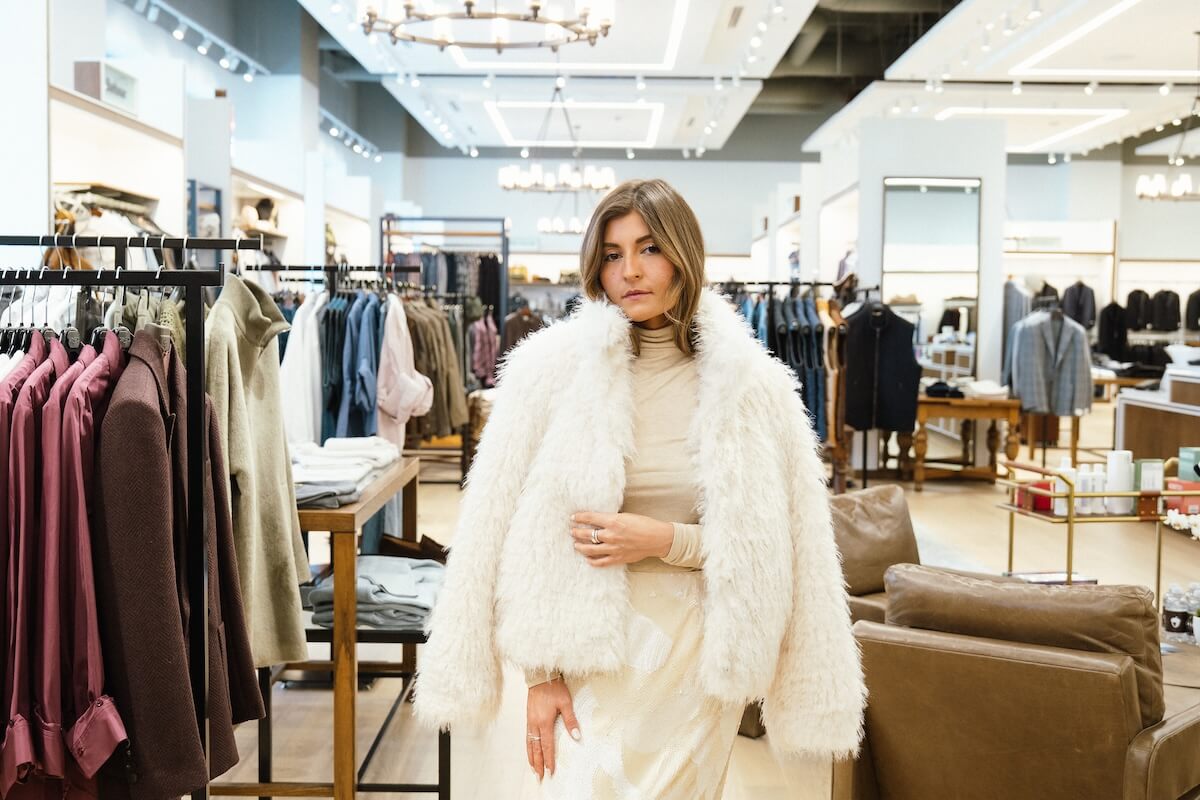 A woman wearing a white faux fur coat stands in a clothing store, browsing racks of clothes and displays as she updates her winter wardrobe.