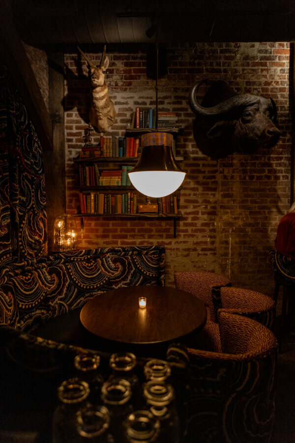 Oxford's New Must-Visit Cocktail Bar: Miscellanea Spirit House - 4 A dimly lit, cozy Oxford cocktail bar with a round table, patterned chairs, brick walls, bookshelves filled with miscellanea, animal head mounts, and a hanging light above the table.