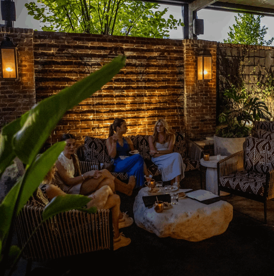 Four women sit and talk amidst outdoor miscellanea on a dimly lit patio with brick walls, plants, lanterns, and a stone table at dusk.