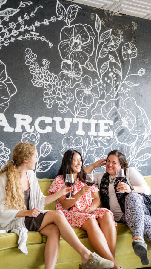 8 Mississippi Destinations Worth the Drive - 4 Three women sit on a yellow couch, smiling and holding glasses of red wine in front of a floral muralβcreating memorable moments at one of the vibrant Mississippi destinations.