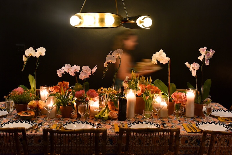 Happy Everything! Laura Johnson’s Joyful Approach to Home & Hosting - 6 A dining table set with floral arrangements, candles, plates, and glasses; a person in the background carries a plate under warm lighting—perfect for happy everything moments and effortless home hosting.