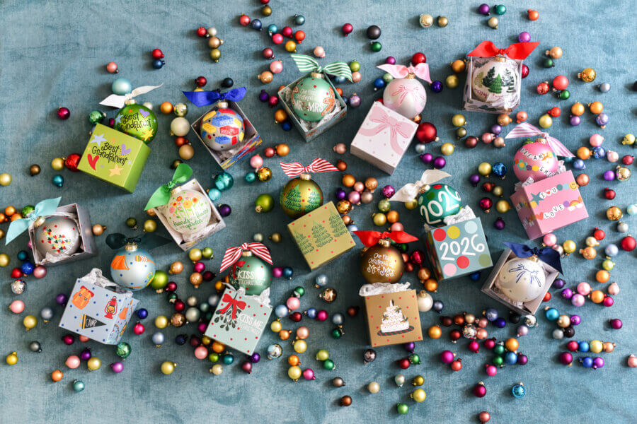 Happy Everything! Laura Johnson’s Joyful Approach to Home & Hosting - 11 Colorful decorated Christmas ornaments in gift boxes are arranged on a blue surface, surrounded by scattered multicolored candy-like balls—a festive touch perfect for home hosting from Happy Everything by Laura Johnson.