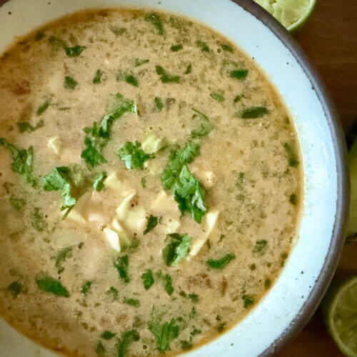 Leftover Chicken Fajita Soup: The Ideal Lazy Girl Recipe - 3 A bowl of creamy Chicken Fajita Soup garnished with chopped cilantro, with halved limes and an avocado half on the wooden table beside it.