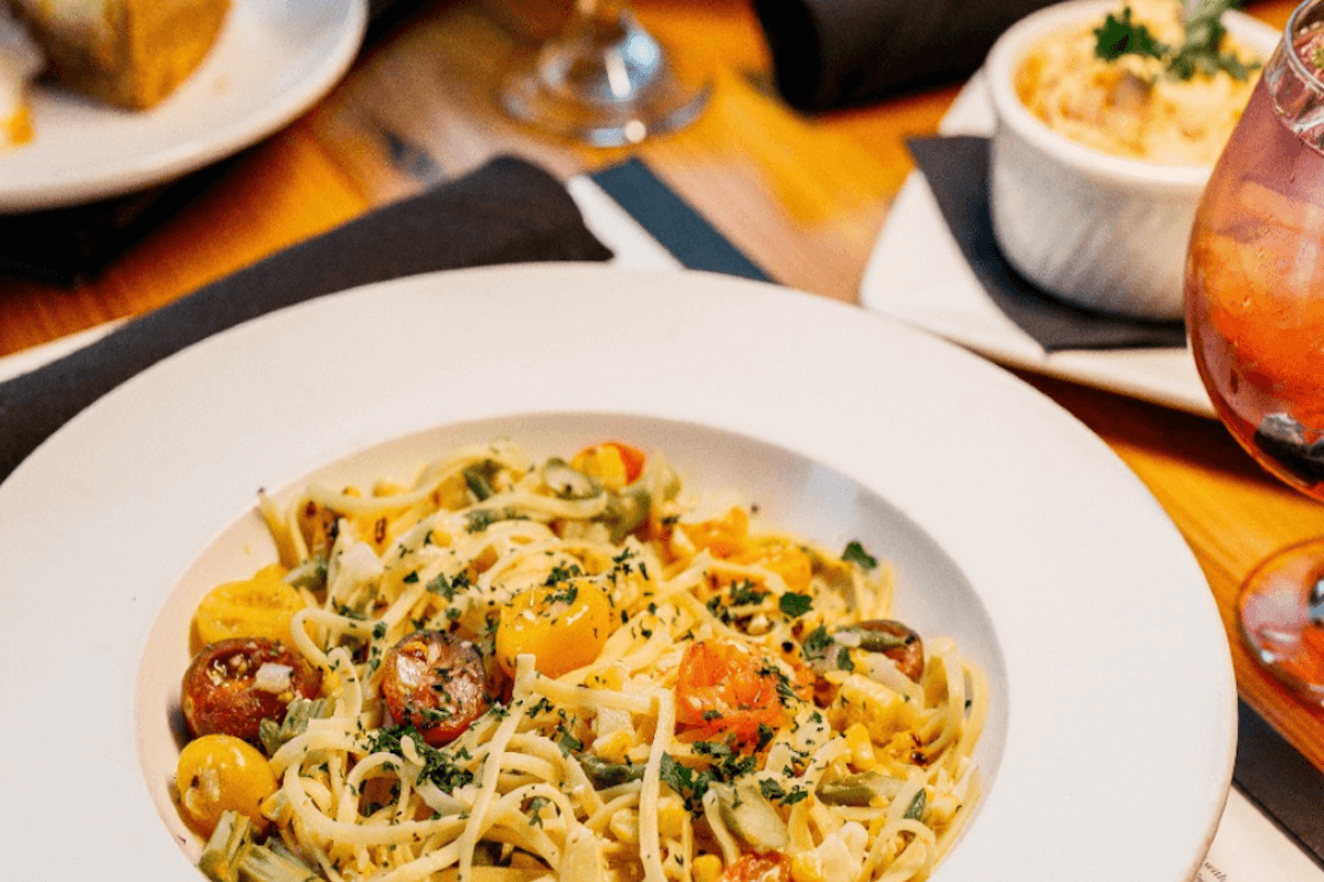 A white plate of pasta with cherry tomatoes and herbs sits on a wooden table, accompanied by a drink and other dishes—perfect for those seeking Memphis restaurants open on Christmas.