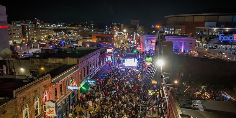 Where to Spend New Year's Eve in Memphis - 2 A large crowd gathers on a brightly lit city street at night for a New Year's Eve celebration, surrounded by buildings, neon signs, and a stadium in the background.