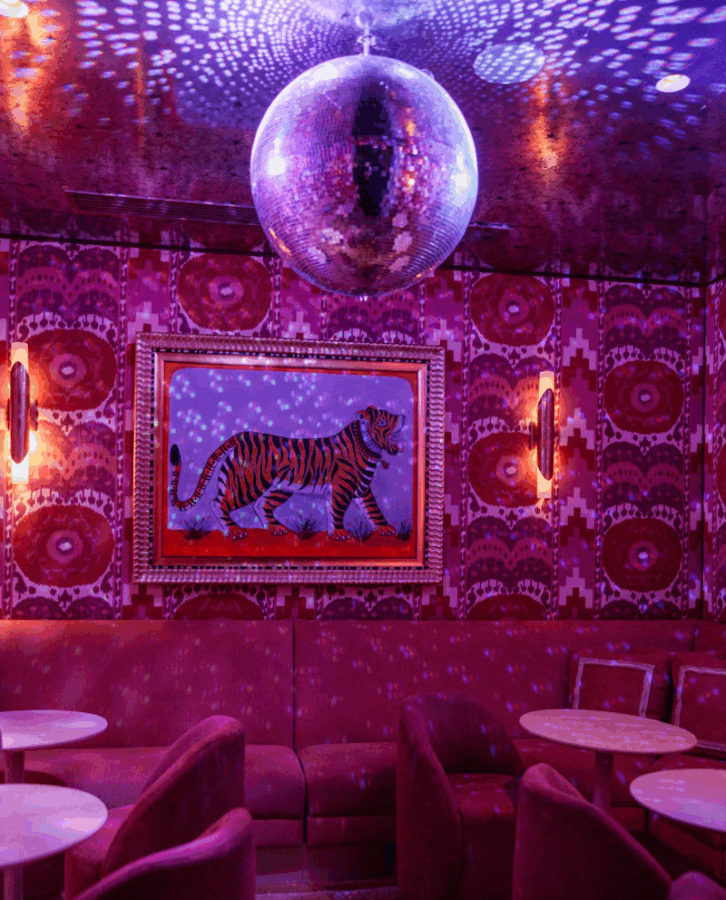 Where to Spend New Year's Eve in Memphis - 3 A room with purple ambient lighting, a disco ball on the ceiling, a framed tiger painting on a patterned wall, and pink upholstered seating around small tablesβperfect for a stylish New Year's Eve celebration in Memphis.
