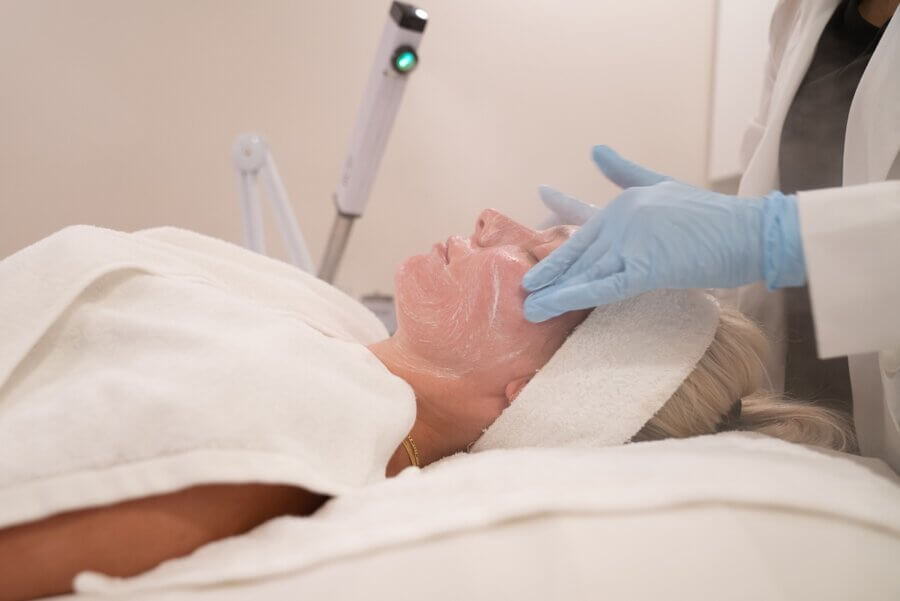 15 Decadent Nashville Spa Treatments to Gift - 3 A person lies on a treatment bed with a towel and headband while a professional wearing blue gloves applies a facial mask in a spa or clinic setting.