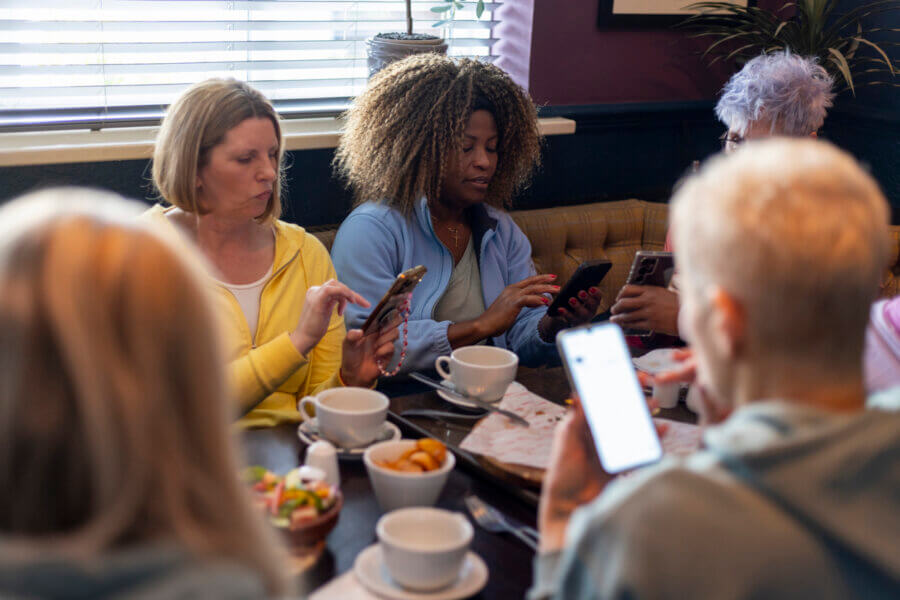 Let's Get Radical: Look People in the Eye and Smile - 3 Five people sit around a table at a cafe, looking at their smartphones. Let's get radical—smile and look people in the eye instead of your screens. Cups, bowls, and plates with food are scattered across the table.