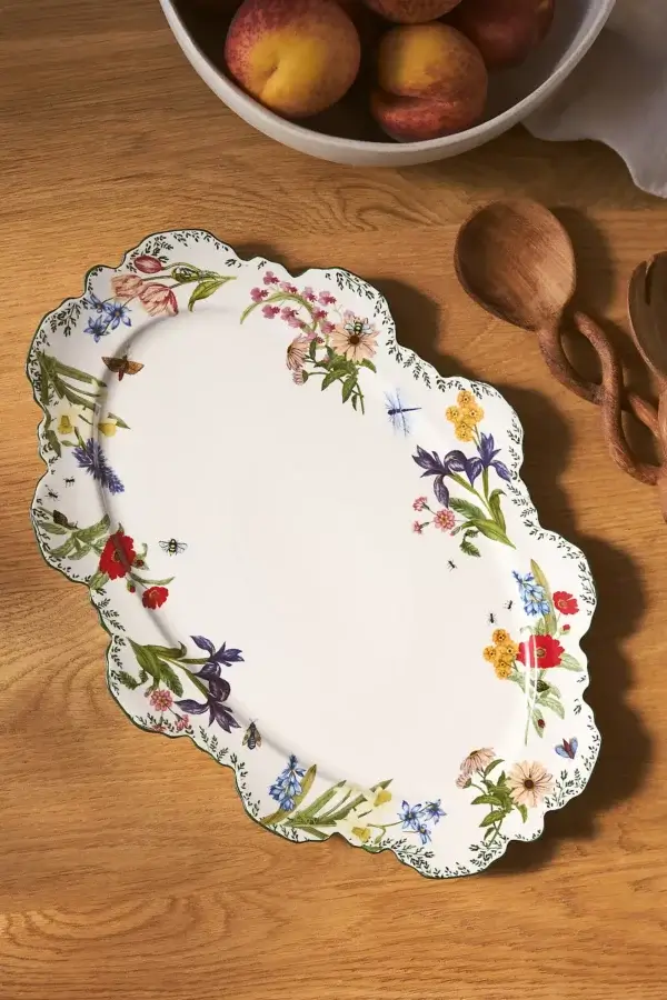 10 Gifts For Bakers Under $100 - 11 An oval ceramic platter with scalloped edges, decorated with colorful floral patterns, sits on a wooden table near a bowl of peaches and wooden utensilsβan ideal pick for baking gifts or thoughtful gifts under $100.