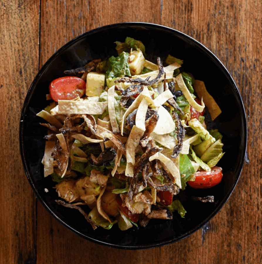 The Best Salads in BHAM: 16 Spots We Love - 12 A black bowl filled with a mixed salad—lettuce, cherry tomatoes, avocado, tortilla strips, and crispy fried onions—sits on a wooden table, showcasing why BHAM Salads are among the best salads you’ll find.