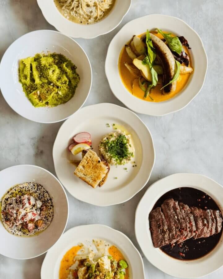 Birmingham's 10 Most Popular Articles of 2025 - 2 Seven white plates with assorted dishes, including sliced beef, pasta, fish, vegetables, grilled sausage with potatoes, and salad, arranged on a light marble tabletop.