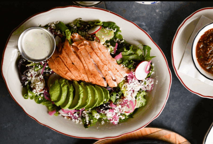 The Best Salads in BHAM: 16 Spots We Love - 9 A plated BHAM Salads creation topped with sliced grilled salmon, avocado, radishes, crumbled cheese, and a side of dressing. A dish of salsa sits nearby.