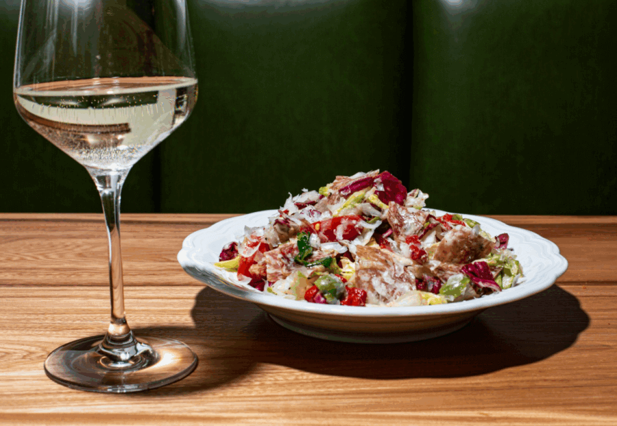 The Best Salads in BHAM: 16 Spots We Love - 10 A glass of white wine and a plate of mixed salad with vegetables and croutons on a wooden table, perfect for enjoying some of the best salads in Birmingham, with a green upholstered background.
