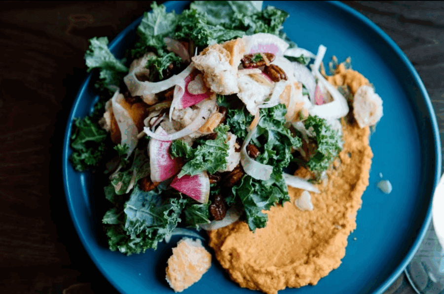 The Best Salads in BHAM: 16 Spots We Love - 13 A plate with a mixed kale salad topped with sliced radish, fennel, pecans, and citrus segments, served beside a smear of orange hummus—one of the best salads in Birmingham from BHAM Salads.