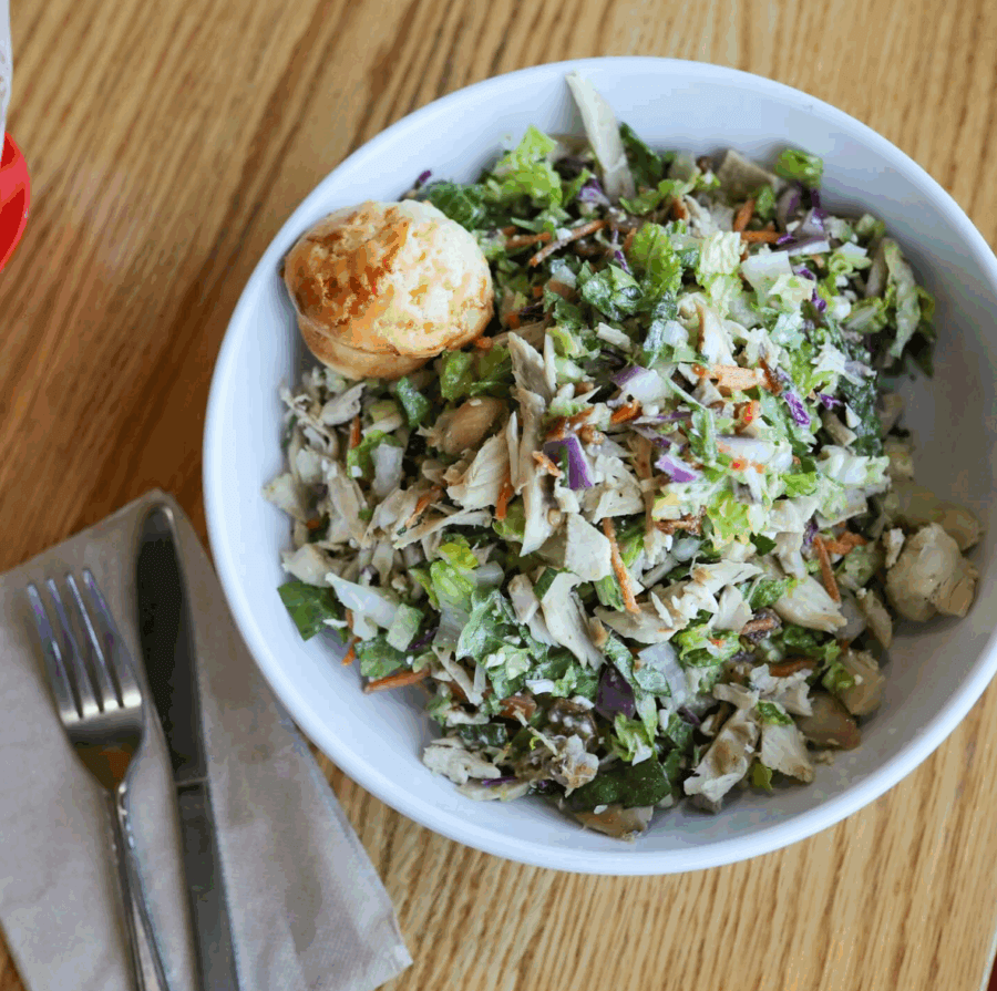 The Best Salads in BHAM: 16 Spots We Love - 16 A white bowl filled with chopped salad and topped with shredded chicken, from BHAM Salads, sits next to a small muffin on a wooden table, with a fork and knife on a napkin.