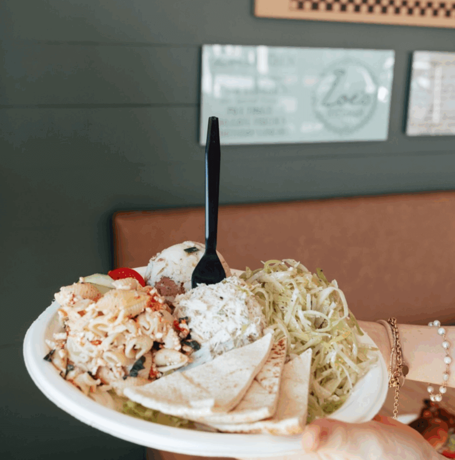 The Best Salads in BHAM: 16 Spots We Love - 17 A hand holds a plate with pasta salad, chicken salad, cole slaw, pita bread slices, and a plastic fork against a green wall background—perfect for anyone searching for the best salads or classic Birmingham salads.