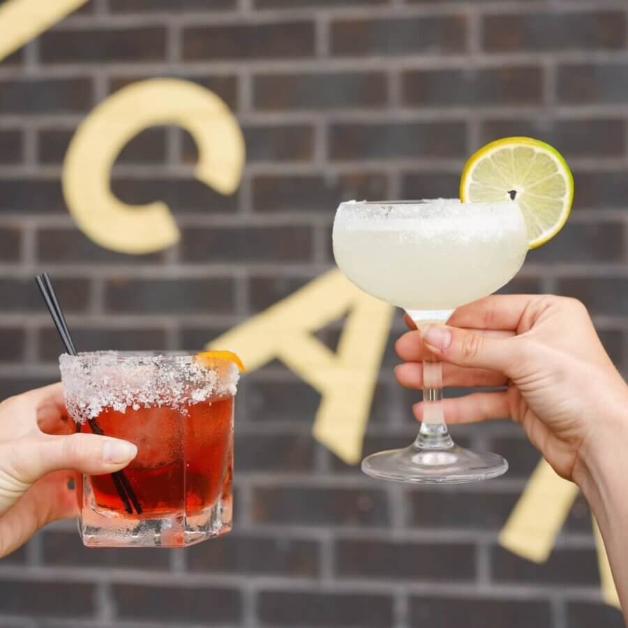 Birmingham's 10 Most Popular Articles of 2025 - 5 Two hands hold cocktails in front of a brick wall in Birmingham; one glass has a red drink with an orange slice, the other has a pale drink with a salted rim and lemon garnish—perfect for those seeking popular articles 2025 inspiration.