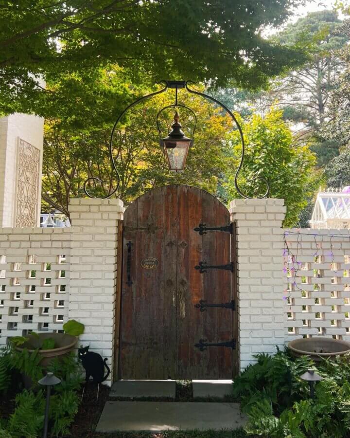 Birmingham's Haven for Hand-Picked Architectural Treasures - 4 A wooden arched gate with metal hinges set in a white brick wall, topped by a hanging Bevolo Lantern, surrounded by greenery and ferns—a charming display of architectural antiques.
