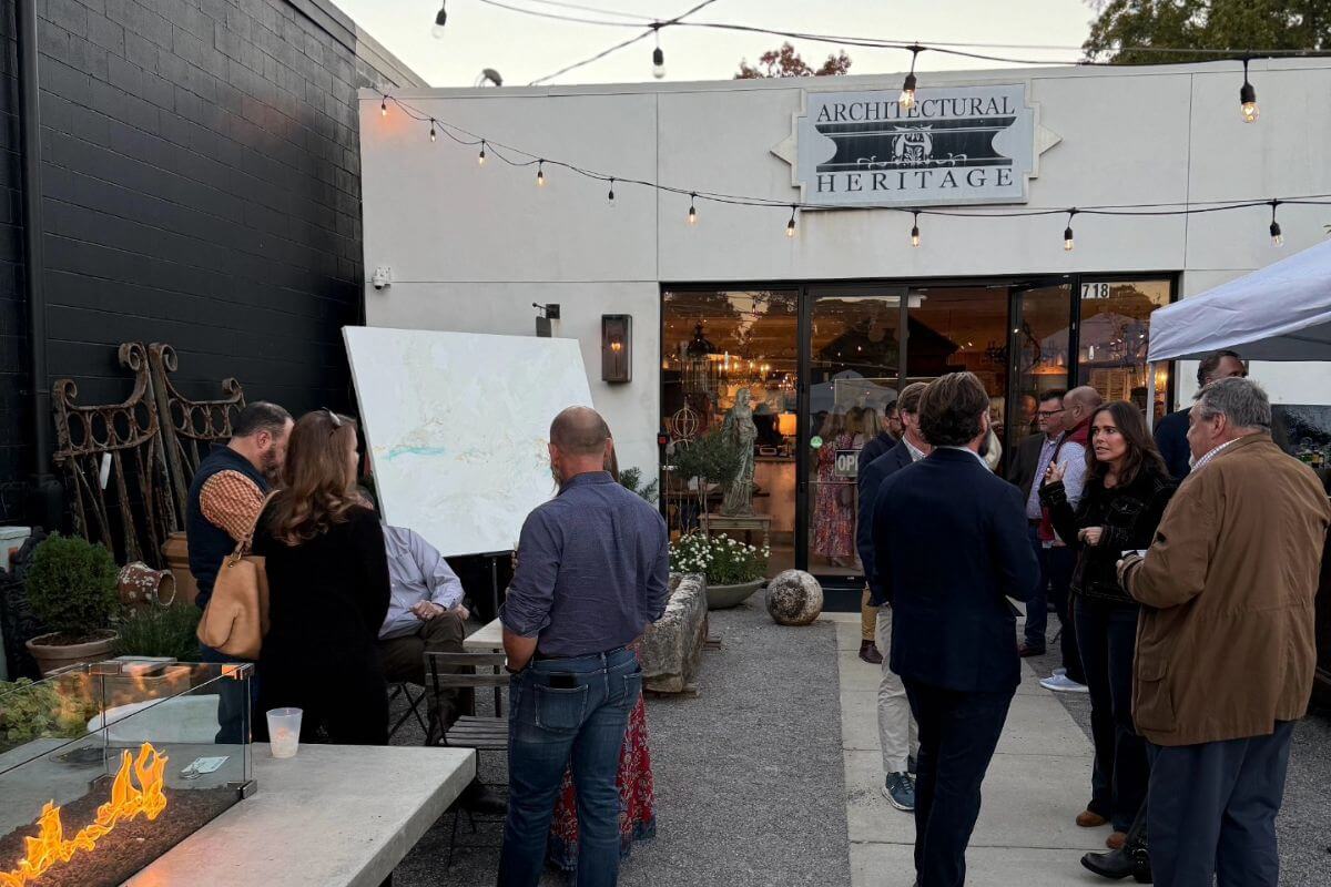 People gather outside Architectural Heritage, conversing around a fire pit and a large display board amid glowing Bevolo lanterns and architectural antiques during an evening event with string lights overhead.