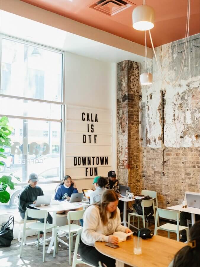 She's Behind One of BHAM’s Most Beloved Coffee Shops - 8 People work and read at tables inside Cala Coffee, a modern BHAM coffee shop with exposed brick walls, large windows, and a sign that reads "CALA IS DTF DOWNTOWN FUN.