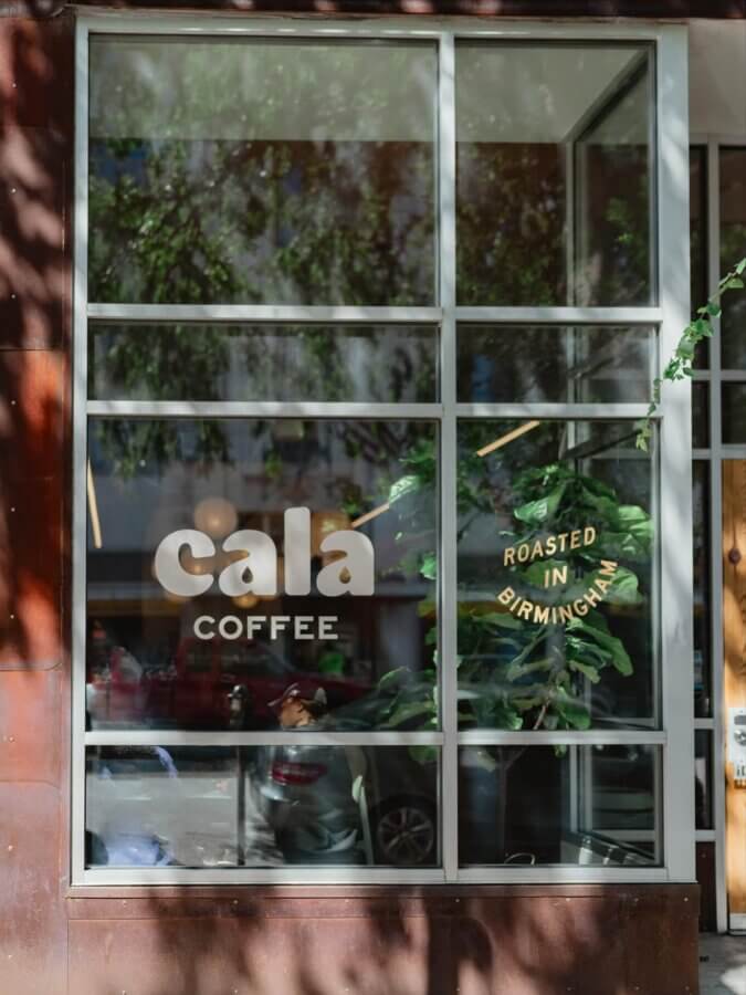 She's Behind One of BHAM’s Most Beloved Coffee Shops - 4 Storefront window with the text "cala COFFEE" and "Roasted in Birmingham" displayed, reflecting trees and parked cars—a true BHAM coffee shop destination.