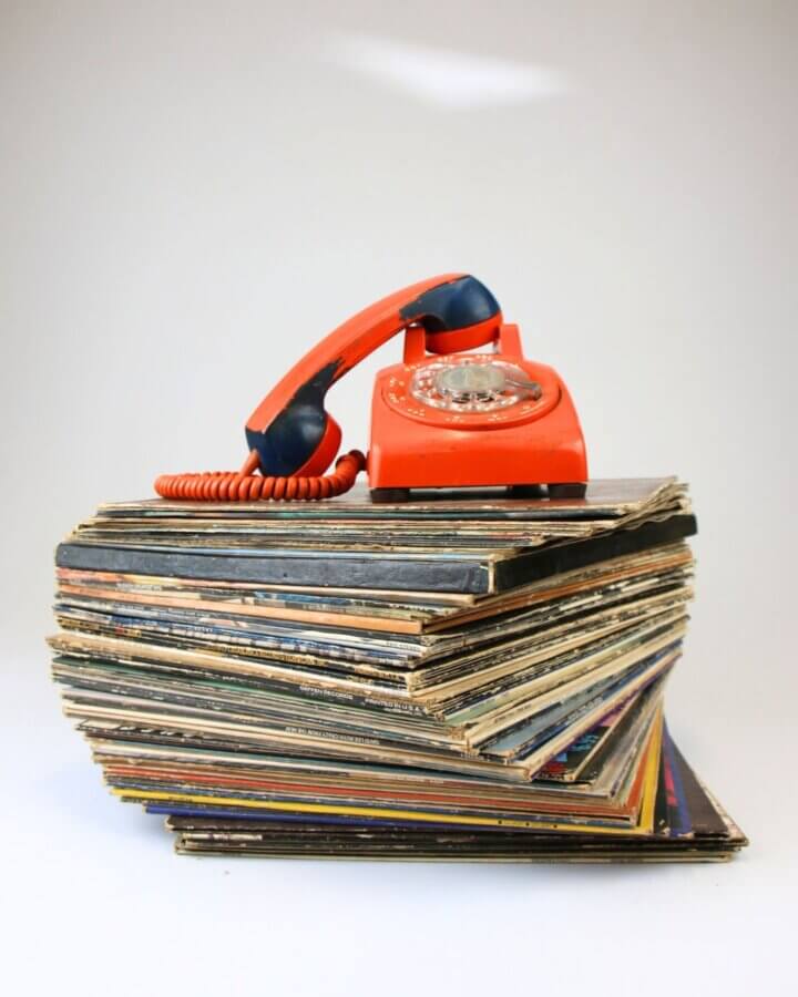 Stop Donating These 5 Items — They May Be Worth Serious Cash - 2 A red rotary telephone rests atop a large, slightly leaning stack of valuable items—vinyl records—against a plain white background.