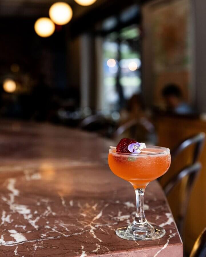 She's Behind La Fête, BHAM's MICHELIN Recognized French Bistro - 7 A coupe glass with an orange cocktail garnished with a slice of strawberry and a small flower sits on a marble bar counter in Birmingham’s La Fête.