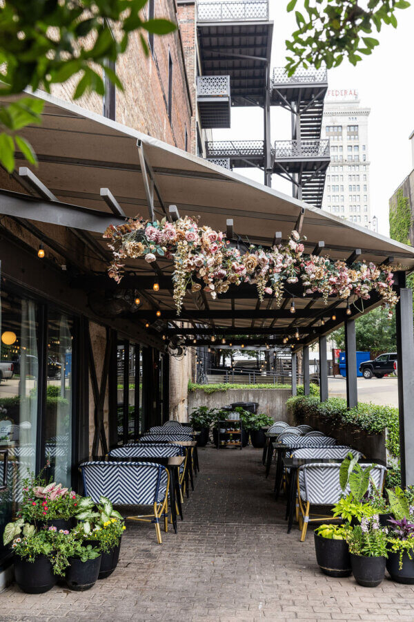 She's Behind La Fête, BHAM's MICHELIN Recognized French Bistro - 4 La Fete's outdoor patio with black-and-white chairs, tables, potted plants, and a floral arrangement hanging from the pergola—creating an inviting spot to relax after donating items worth serious cash—set beside a brick building and city street.