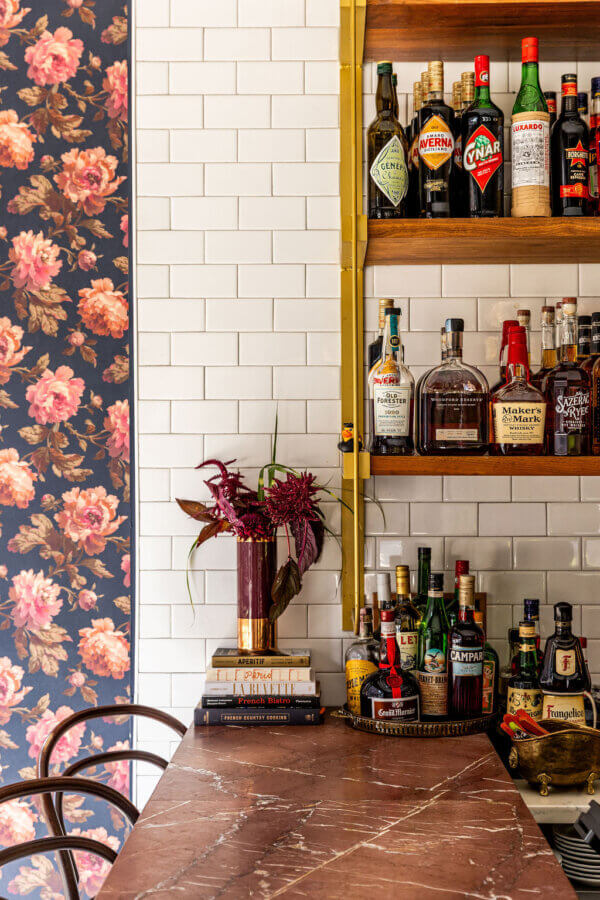 She's Behind La Fête, BHAM's MICHELIN Recognized French Bistro - 5 Marble bar counter with assorted liquor bottles on wooden shelves, a floral wallpaper on the left, and a vase with red flowers on a stack of valuable items.