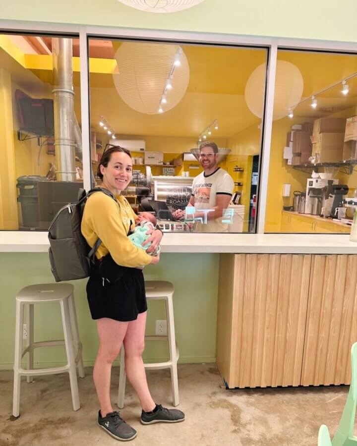 She's Behind One of BHAM’s Most Beloved Coffee Shops - 7 A woman holding a baby stands by a counter at a café, smiling at a man behind the glass who is preparing drinks.