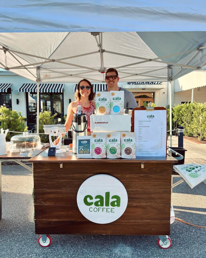 She's Behind One of BHAM’s Most Beloved Coffee Shops - 5 Mel and Josh Cosio stand behind a wooden coffee cart labeled "Cala Coffee," with a menu, bags of coffee, pastries, and a coffee machine visible under a canopy at an outdoor market.