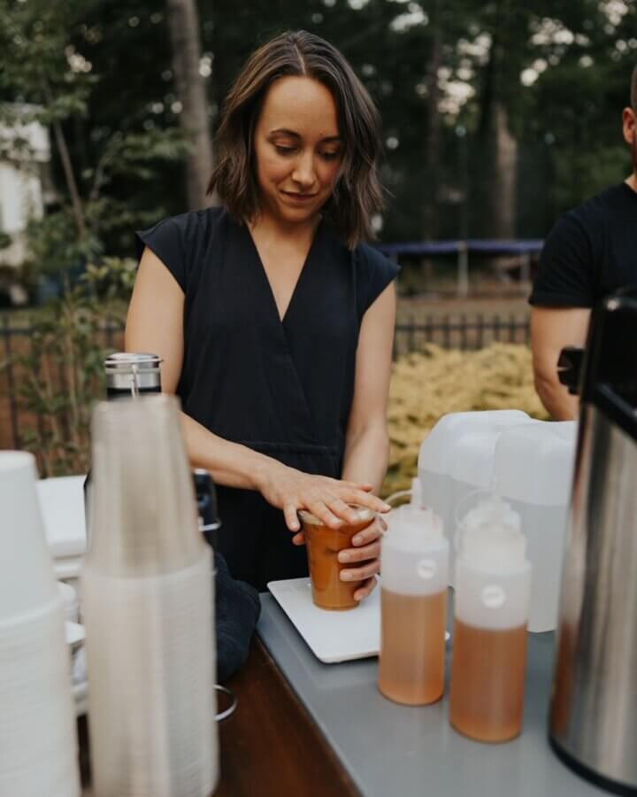 She's Behind One of BHAM’s Most Beloved Coffee Shops - 2 Mel Cosio in a black dress closes a to-go cup at an outdoor drink station with cups, bottles, and dispensers on the table.