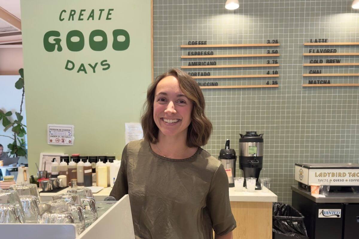 Mel Cosio stands and smiles behind the counter at Cala Coffee, with a menu board and the phrase “CREATE GOOD DAYS” visible on the wall.