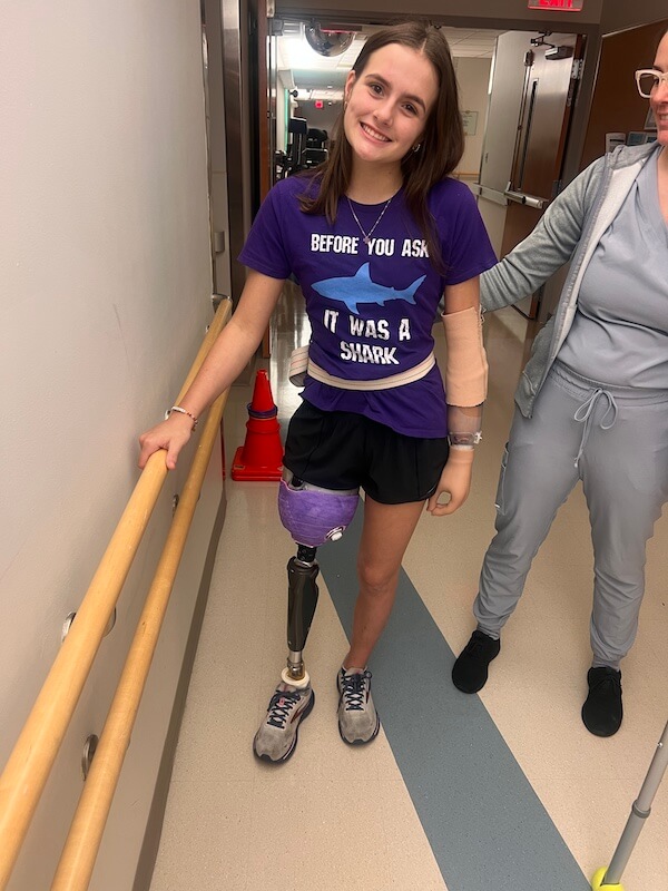 Meet 17-Year-Old Shark Attack Survivor Lulu Gribbin - 3 17-year-old shark attack survivor Lulu Gribbin stands in a hallway, smiling in her purple "Before you ask, it was a shark" shirt. She wears a prosthetic leg and arm brace as an adult stands beside her.