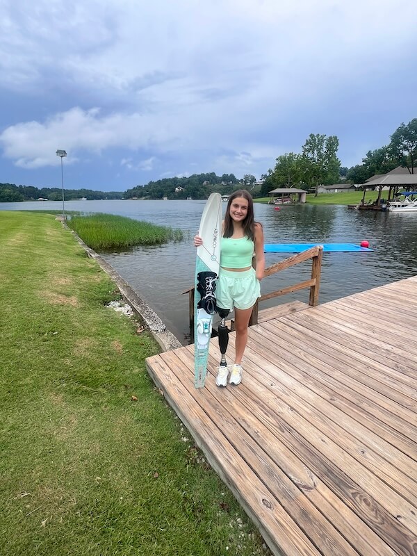 Meet 17-Year-Old Shark Attack Survivor Lulu Gribbin - 5 17-year-old shark attack survivor Lulu Gribbin stands on a wooden dock by a lake, holding a water ski, with grassy shoreline and boats visible in the background.