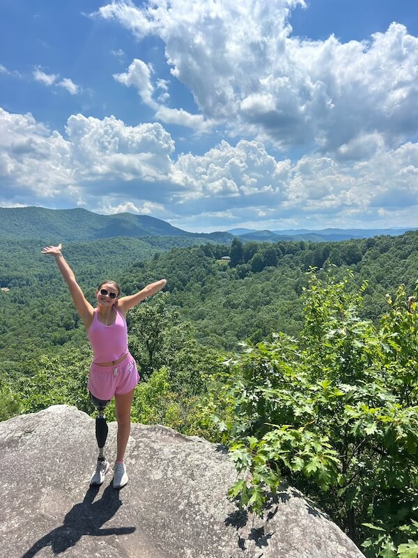 Meet 17-Year-Old Shark Attack Survivor Lulu Gribbin - 6 A survivor of a shark attack, a woman with a prosthetic leg stands on a rocky overlook, smiling with arms raised, green forest and mountains beneath a partly cloudy sky in the background.